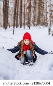 A teenage girl in bright clothes sits on the snow and smiles. Walk through the coniferous forest during the cold season. Knitted warm accessories in trendy colors. Vertical photo of children.