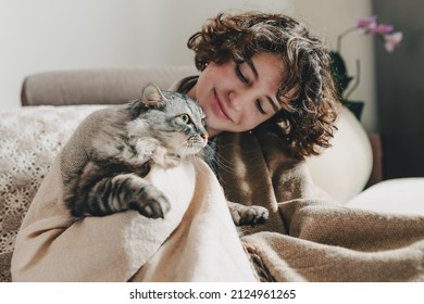 teenage girl with brown curly hair sits on sofa, wrapped in blanket, smiles sweetly, hugs beloved domestic gray cat, which purrs. Showing love for pets while in cozy home, real people