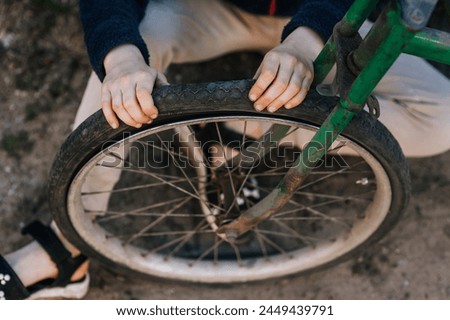 A teenage girl, a child, sits near an old retro bicycle with a broken, punctured wheel outdoors, pressing her hands on the tire. Close-up photography, portrait, lifestyle.