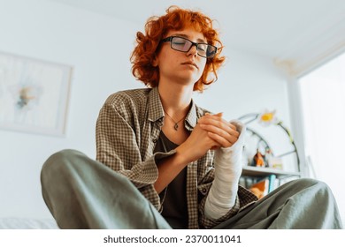 teenage girl, with curly red hair, wearing glasses, with broken arm in cast, sits on bed in room, thoughtfully, dreams or prays - Shutterstock ID 2370011041