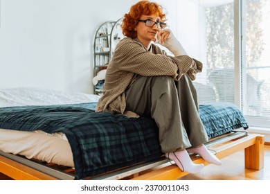 teenage girl, with curly red hair, wearing glasses, with broken arm in cast, sits on bed in room, thoughtfully, dreams or prays - Shutterstock ID 2370011043