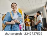 teenage girl with digital tablet looking at camera in school hallway, blurred background, students