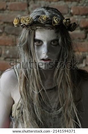 A teenage girl dresses as a Halloween horror figure. 
Her face reveals an astonished look as she peers to
the camera. She wears a thorny crown on her head. 
