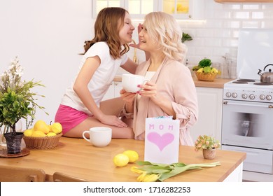 Teenage girl drinking tea with mother at Mother s Day. Daughter sitting on table and whispering something on ear of her mother. Family, holiday, birthday concept