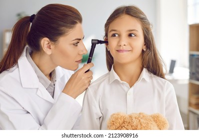 Teenage girl is examined by otolaryngologist during scheduled medical examination at hospital. Close-up of female ENT doctor with otoscope examines ear of smiling child who has funny facial expression