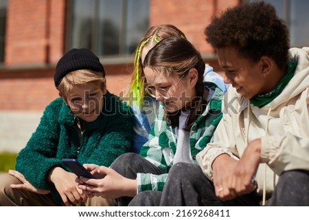 Teenage girl in eyeglasses communicating online on mobile phone while sitting outdoors with her friends