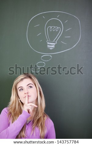 Teenage girl with finger on lips looking up while standing next to bulb drawn in thought bubble against chalkboard