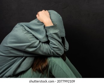 Teenage girl in a green sweatshirt covers her face with her hands on a dark background. The girl sits alone and is afraid. Adolescent psychology concept. Emotions.