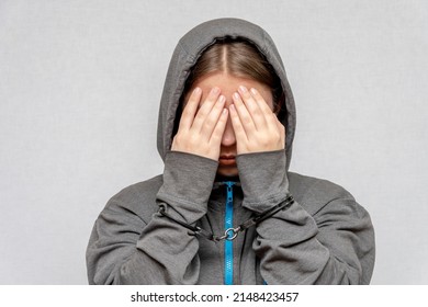 A Teenage Girl Is Handcuffed, Hiding Her Face, In A Hoodie, On A Gray Background. Juvenile Delinquent, Criminal Liability Of Minors. Members Of Youth Criminal Groups And Gangs.