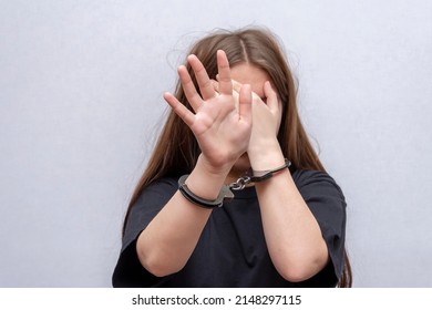 A Teenage Girl In Handcuffs Hides Her Face On A Gray Background. Juvenile Delinquent In A Black T-shirt, Criminal Liability Of Minors.