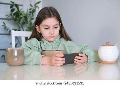 La adolescente tiene desayuno en la cocina, utiliza el teléfono móvil mientras desayuna en la mesa. Muchos niños no quieren comer a menos que estén viendo un dispositivo móvil o TV.
