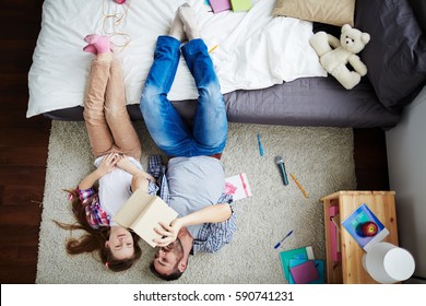 Teenage Girl And Her Middle-aged Father Lying On Messy Bedroom Floor And Watching Photo Book, Feet Up On Bed