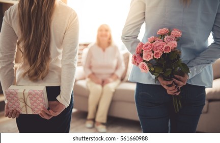 Teenage girl and her mom are hiding flowers and a gift box for their beautiful granny behind backs while grandma is sitting on couch at home