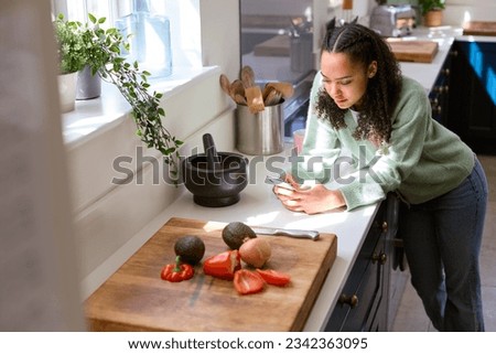 Teenage Girl At Home In Kitchen With Ingredients Looking At Recipe On Mobile Phone