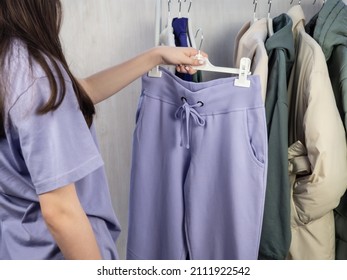 A teenage girl in a lilac T-shirt chooses what to wear. Lilac sweatpants, a white jacket and a sweatshirt hang on a hanger. Women's, youth clothes.