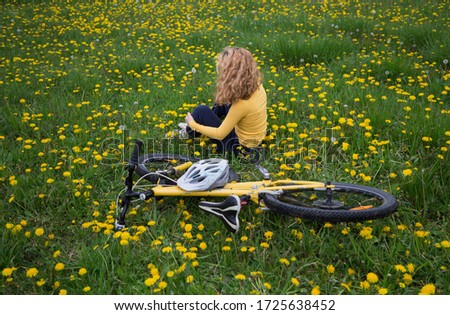 teenage girl with magnificent blond hair sits on green grass among yellow dandelions, a bicycle lies nearby. outdoor bike ride, harmony, energy of nature, relaxation, freedom. Active healthy lifestyle