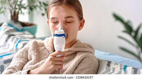 Teenage Girl Makes Inhalation With A Nebulizer Equipment. Sick Child Holding Inhalator In Hand And Breathes Through An Inhaler At Home. Physical Therapy For Cold, Flu And Broncial Asthma