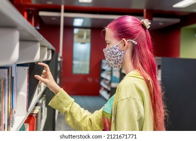 Teenage Girl With Ombre Pink Purple Colored Hair Wearing Face Mask Choosing Books At The Local Library During The COVID-19 Pandemic. School Library