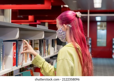 Teenage Girl With Ombre Pink Purple Colored Hair Wearing Face Mask Choosing Books At The Local Library During The COVID-19 Pandemic. School Library