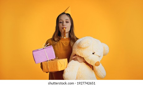 Teenage Girl In Party Cap Blowing Party Horn While Holding Presents And Soft Toy Isolated On Yellow