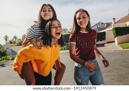 Teenage girl piggy riding on her friend walking in the street on a sunny day. Three smiling girls having fun walking in the street on a sunny day.