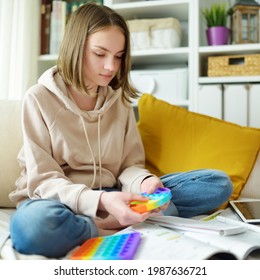 Teenage Girl Playing With Rainbow Pop-it Fidget Toy While Studying At Home. Teen Kid With Trendy Stress And Anxiety Relief Fidgeting Game. Popping The Dimples Of Sensory Silicone Toy.