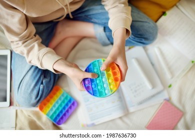 Teenage Girl Playing With Rainbow Pop-it Fidget Toy While Studying At Home. Teen Kid With Trendy Stress And Anxiety Relief Fidgeting Game. Popping The Dimples Of Sensory Silicone Toy.