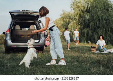 Teenage Girl Playing with West Highland White Terrier Dog on Background of Her Family Having Fun Time Outdoors on Road Trip by Minivan Car