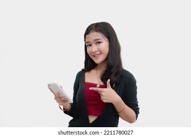 A Teenage Girl Pointing At Her Mobile Phone As Scrolls Through Her Social Media Accounts. Studio Photo With Plain White Backdrop.