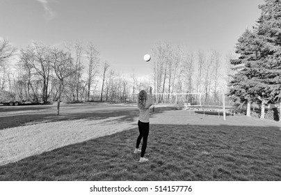 A Teenage Girl Practicing Serving Volleyball Over Net In Backyard In Black And White