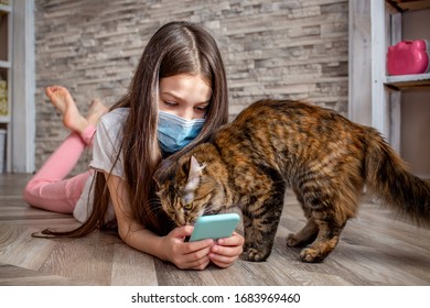 Teenage Girl In Protective Mask On Her Face And Her Cat Lies On The Floor With Smartphone. Coronavirus Quarantine In Europe