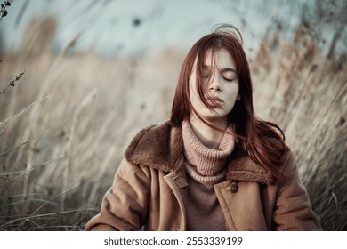 Teenage girl with red hair and closed eyes sitting outdoors in tall dry grass, wearing a warm brown coat and pink sweater, conveying a peaceful and reflective mood in an autumn field. - Powered by Shutterstock