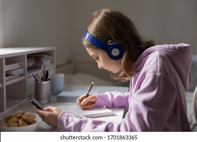 Teenage Girl School Pupil Wearing Headphones Holding Smart Phone, Studying, Making Notes. Focused Teen Student Doing Homework Using Mobile App For Remote Education Listening Audio Lesson At Home.