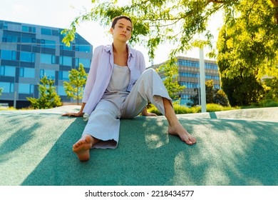 Teenage Girl With Short Hairstyle, Unisex Clothing, Sits On Artificial Turf, Barefoot, Against Blurred Background Modern Architecture, Residential Real Estate Or Glass Facades. Adolescence, Portrait 