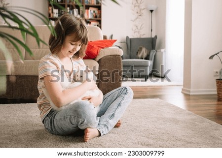 A teenage girl sits on the floor in the living room and plays with her beloved kitten. The child hugs and strokes the pet. Children and cats.