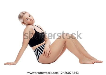 A teenage girl sits in a sports swimsuit. A thoughtful blonde with freckles on her face in a black top and striped panties. Isolated on a white background.