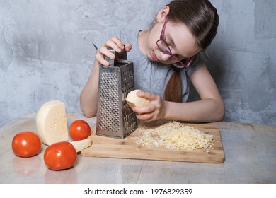 A teenage girl sits at the table and grates cheese on a grater on a wooden board. Cheese and tomatoes are on the table next to it. - Powered by Shutterstock