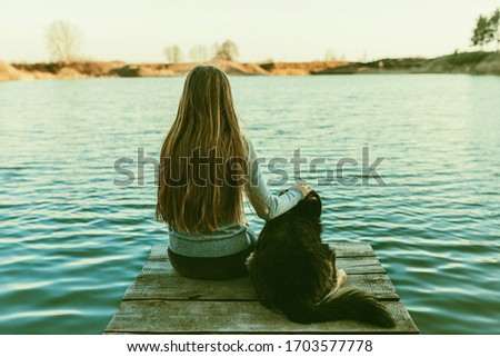 A teenage girl is sitting with a dog on the bridge and looking at the lake. Friendship between human and dog. Pets and animals concept