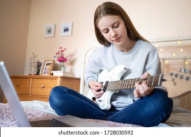 Teenage Girl Sitting On Bed Learning To Play Electric Guitar With Online Lesson On Laptop Computer