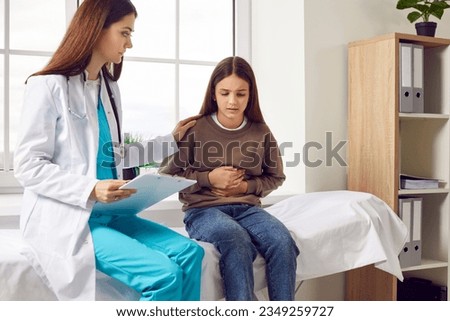 Teenage girl sitting on the couch in the doctor's office and pointing to her stomach to paediatrician during medical examination in clinic. Child doctor listening to the patient's complaints.