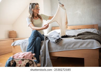 A teenage girl sitting on her bed, trying on different outfits. She holds up various clothes, contemplating her choices as she decides what to wear for the day. - Powered by Shutterstock