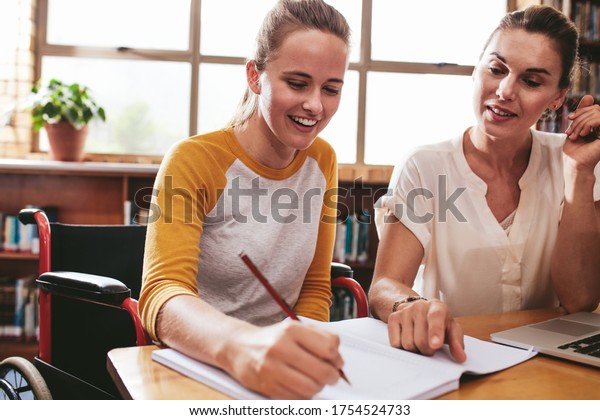 Teenage girl sitting in a wheelchair writing in a\
notebook with a female teacher sitting by at school. Disabled\
female student studying with\
tutor.