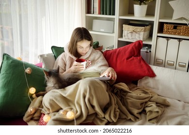 Teenage girl snuggling up on the sofa in a cozy living room at Christmas. Cute child using a tablet at home during winter break. Kid reading an ebook in comfy blanket. Celebrating Xmas at home. - Powered by Shutterstock