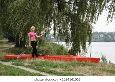 A teenage girl stands near a red kayak and holds an oar in her hand. Concept of physical and mental health of teenagers - Powered by Shutterstock