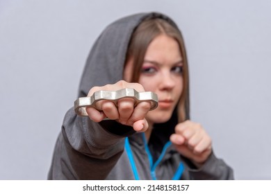 A Teenage Girl Strikes With Brass Knuckles On A Light Background, Wearing A Hoodie With A Hood. Concept: Street Gang Of Hooligans, Infliction Of Grievous Bodily Harm, Threat To Life And Health.