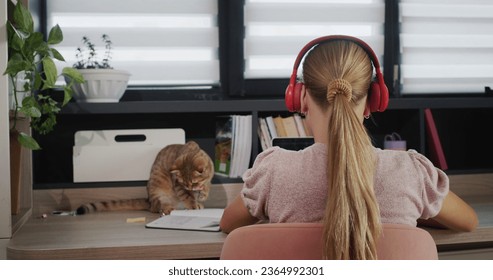 A teenage girl is studying at a laptop at home. She has headphones on, her pet helper sits next to her - Shutterstock ID 2364992301