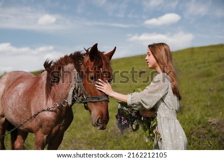 A teenage girl in a summer turquoise dress with a bouquet of lilacs strokes a brown horse in a meadow.