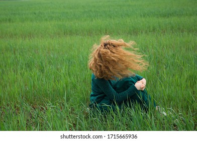 teenage girl in sweater and long emerald-colored skirt sits on ground among field of young grass, her face is covered with hair that flutters in  wind. Harmony, inspiration, relaxation, self-isolation