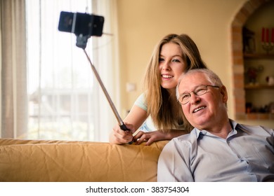 Teenage Girl Taking Photo With Mobile Phone On Selfie Stick Of Herself And Her Grandfather At Home