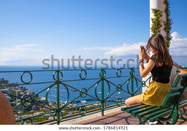 Teenage girl taking\
pictures during summer vacation of fantastic view of sea whith\
smartphone camera. Brunette girl with long hair standing backwards\
and taking the picture